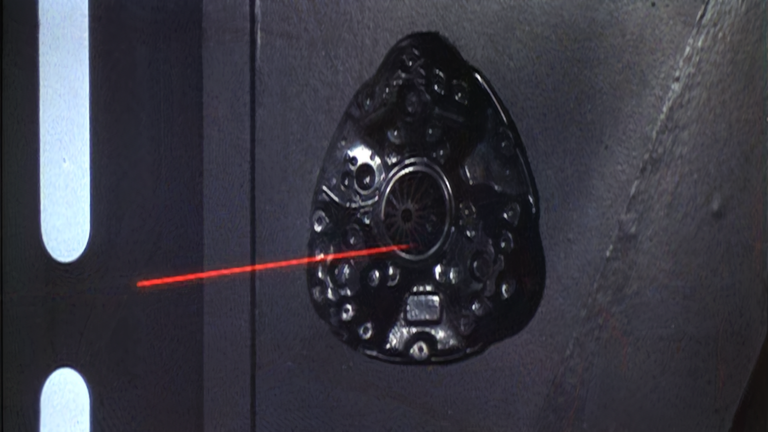 Star Wars still of a red laserbeam about to hit a black, shell looking speaker-like wall fixture